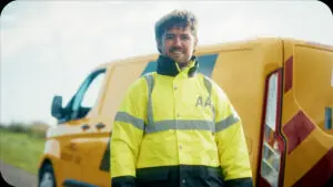 Smiling AA Patrol wearing a high-visibility jacket in front of a yellow roadside-assistance van.
